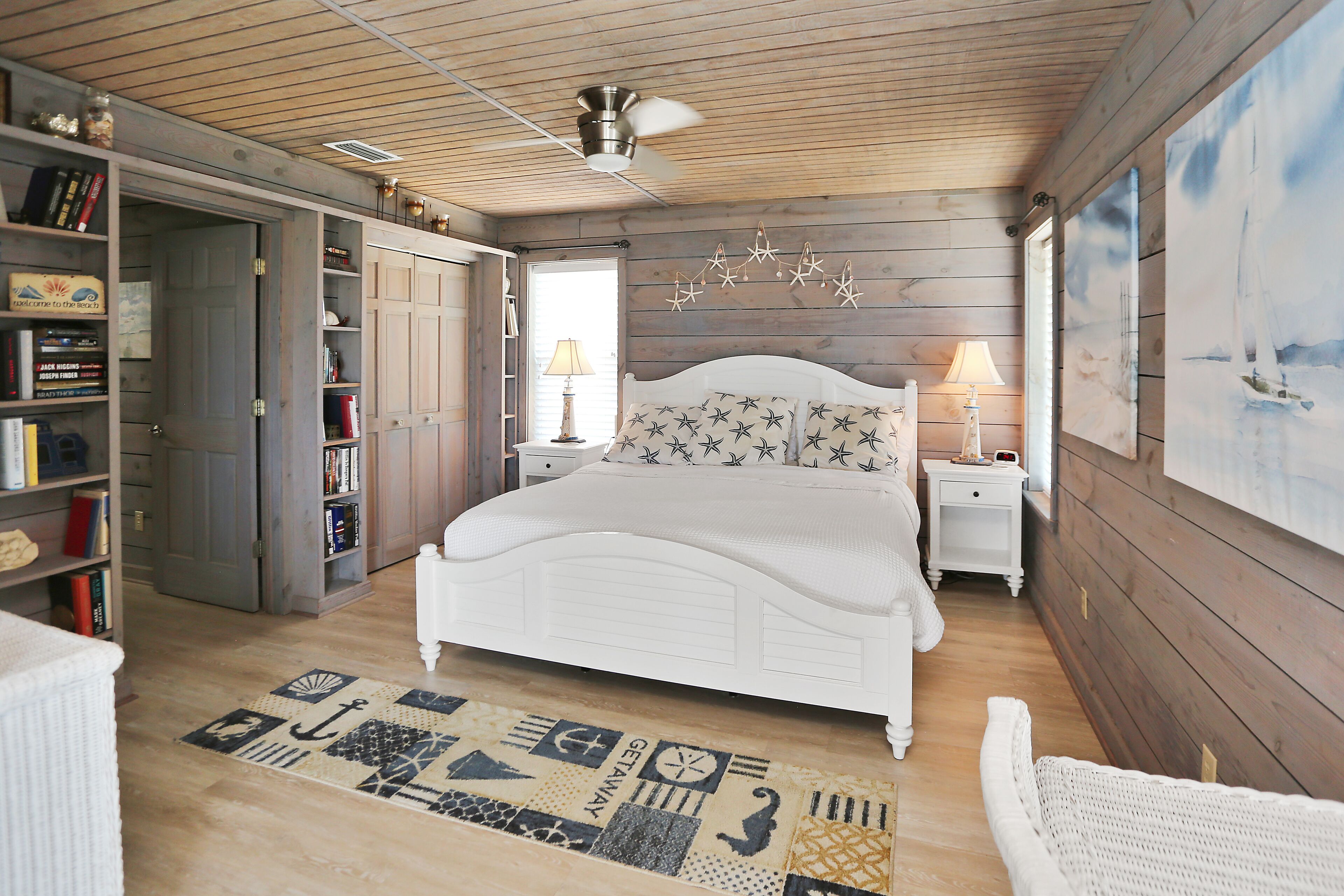 A bedroom with a wooden ceiling, a white bed, a patterned rug, and a sailboat painting on the wall.