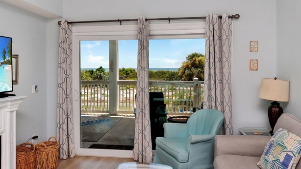Un salon comprenant un canapé, un fauteuil et un téléviseur. Il y a une grande fenêtre avec des rideaux, une cheminée et une vue sur un balcon avec une piscine.