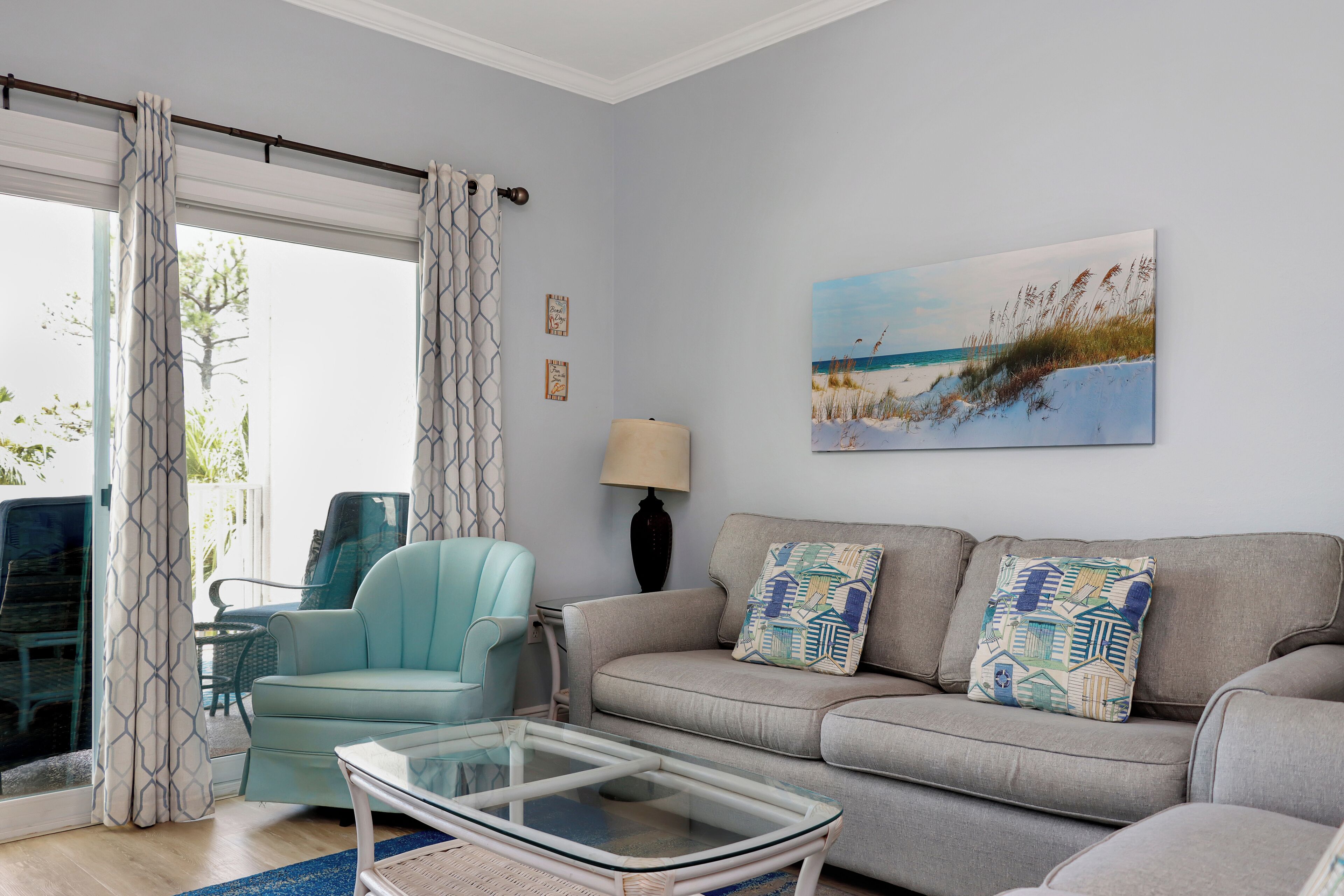 Un salon comprenant un canapé, un fauteuil, une table basse en verre et un tableau représentant une scène de plage.