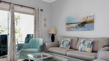 Un salon comprenant un canapé, un fauteuil, une table basse en verre et un tableau représentant une scène de plage.