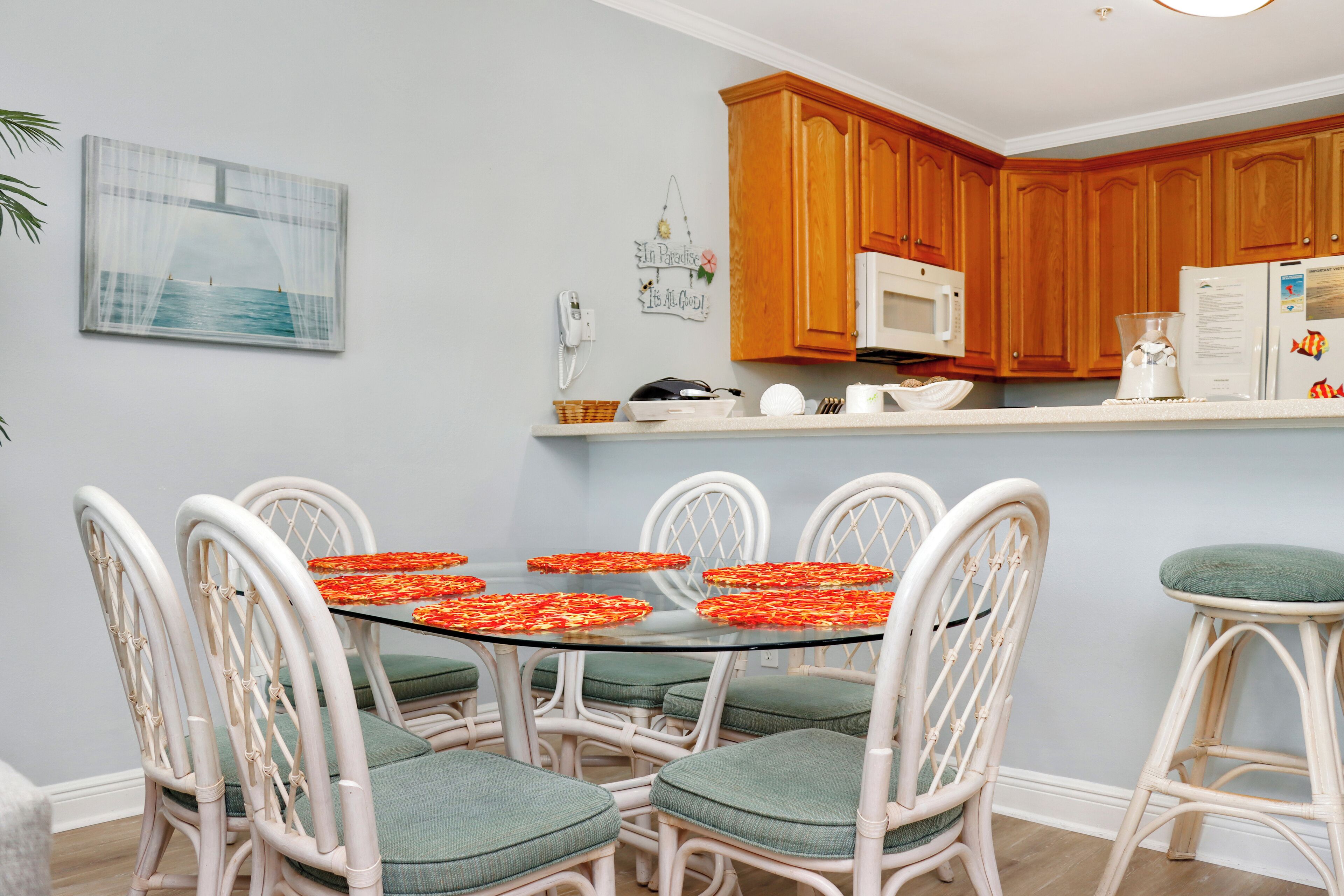 Un comedor con una mesa de vidrio rodeada de sillas blancas con cojines verdes, armarios de madera, un microondas blanco y un refrigerador.