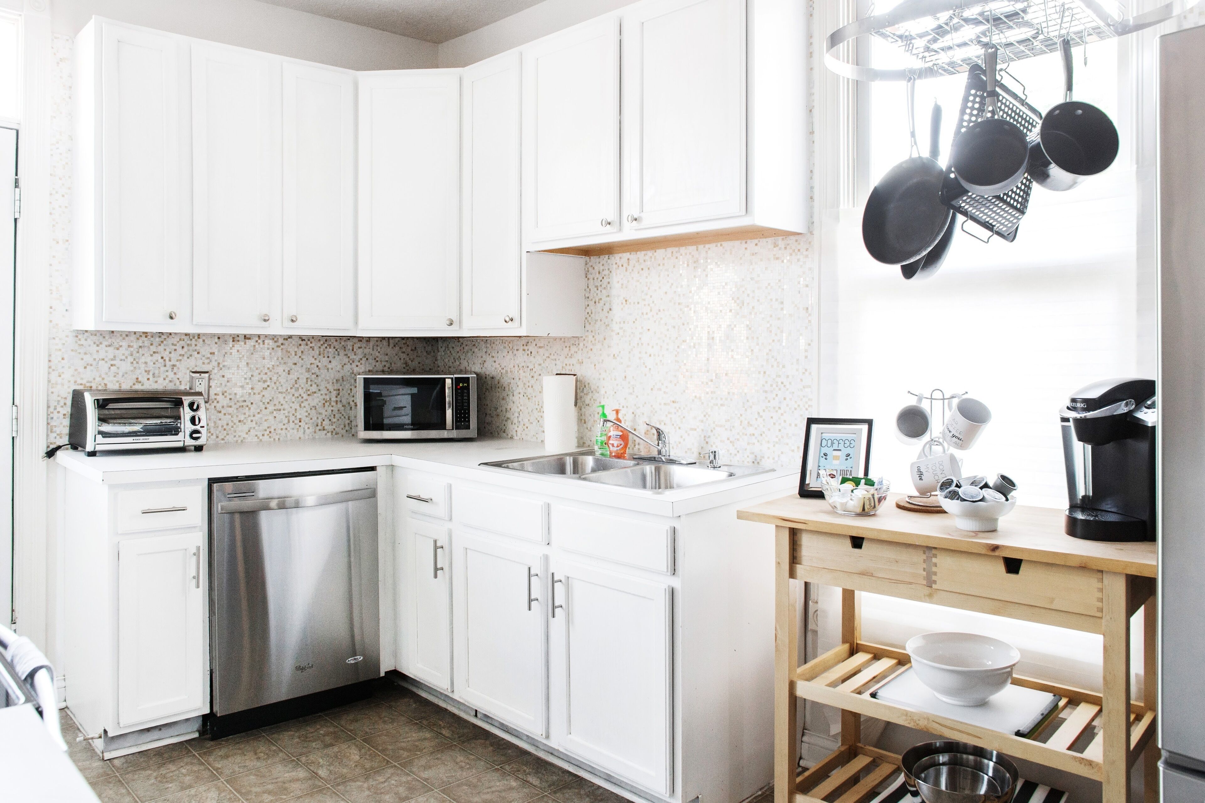 A modern kitchen with white cabinets, a dishwasher, a microwave, a coffee maker, and a wooden table with a bowl.
