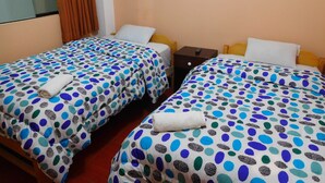Quarto de hotel com duas camas de solteiro, cada uma com colcha de bolinhas e um travesseiro branco. Há uma pequena mesa de cabeceira de madeira entre as camas, com um controle remoto sobre ela.