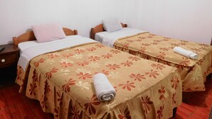Quarto de hotel com duas camas, cada uma com colcha florida e toalha enrolada. Há uma mesa de cabeceira com um controle remoto e uma televisão na parede.