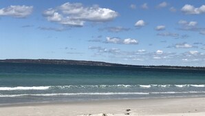A beach with clear blue water, a sandy shore, and a distant landmass with buildings. - Little Lewis - Romantic Getaway (Callala Beach)