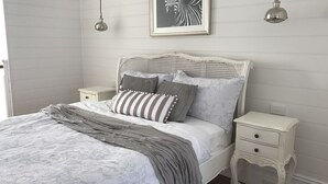 A neatly made bed with a grey and white striped pillow, a grey throw blanket, and a white headboard with a woven pattern. There are two bedside tables with drawers, a framed picture on the wall, and two hanging pendant lights. - Little Lewis - Romantic Getaway (Callala Beach)