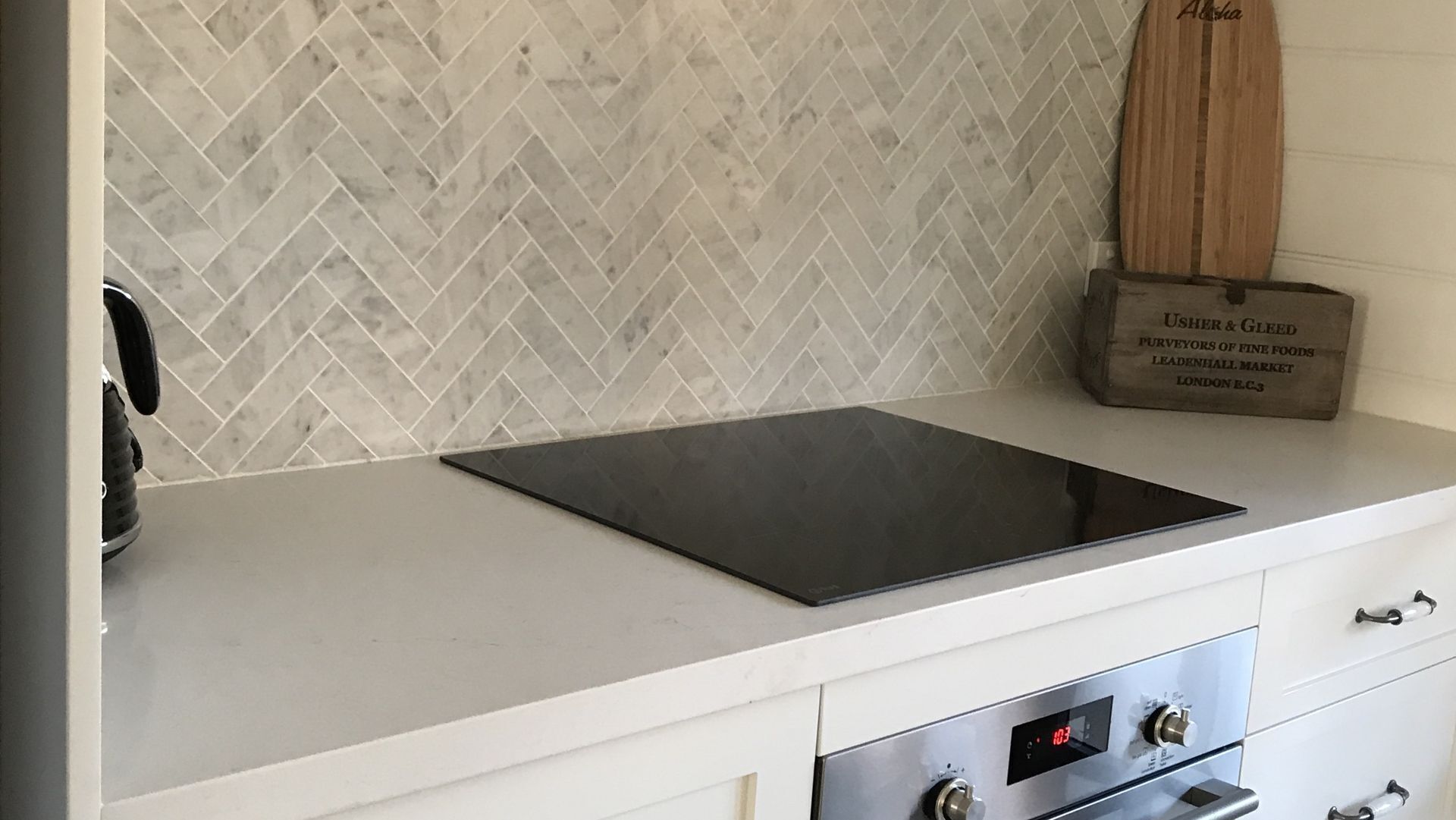 A kitchen with a herringbone-patterned backsplash, a black induction cooktop, and a wooden box labeled "Usher & Gled".