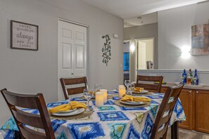 A dining area with a table set for four, chairs, a decorative wall piece, and a kitchen in the background. - Superstition Lakes Condo, A+ Golf View, Comm Htd POOL & SPA; Walk to Mall & Theater (Mesa)