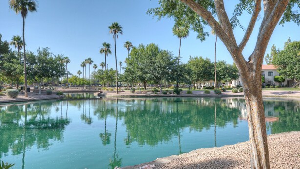 A serene outdoor area with a calm pond, surrounded by palm trees and a building with a red roof. - Superstition Lakes Condo, A+ Golf View, Comm Htd POOL & SPA; Walk to Mall & Theater (Mesa)