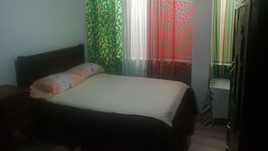Een slaapkamer met een bed, nachtkastje, radiator en gordijnen met een bladmotief.