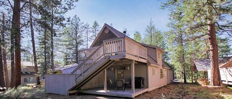 A two-story cabin with a balcony and a covered outdoor seating area, surrounded by tall pine trees.