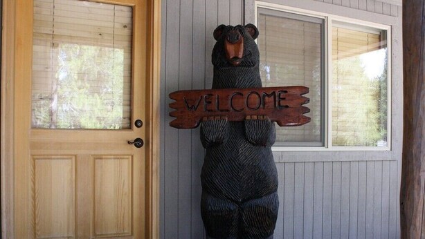 Une sculpture en bois représentant un ours, tenant une pancarte sur laquelle est écrit « BIENVENUE », à l’entrée d’un bâtiment.