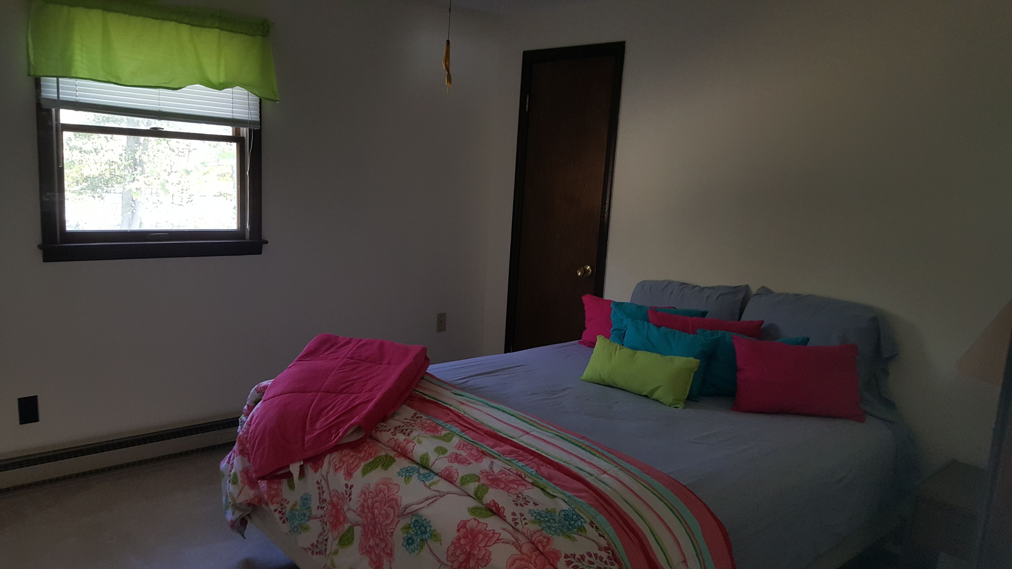 Un dormitorio con cama, ventana con cortinilla verde, puerta y radiador.