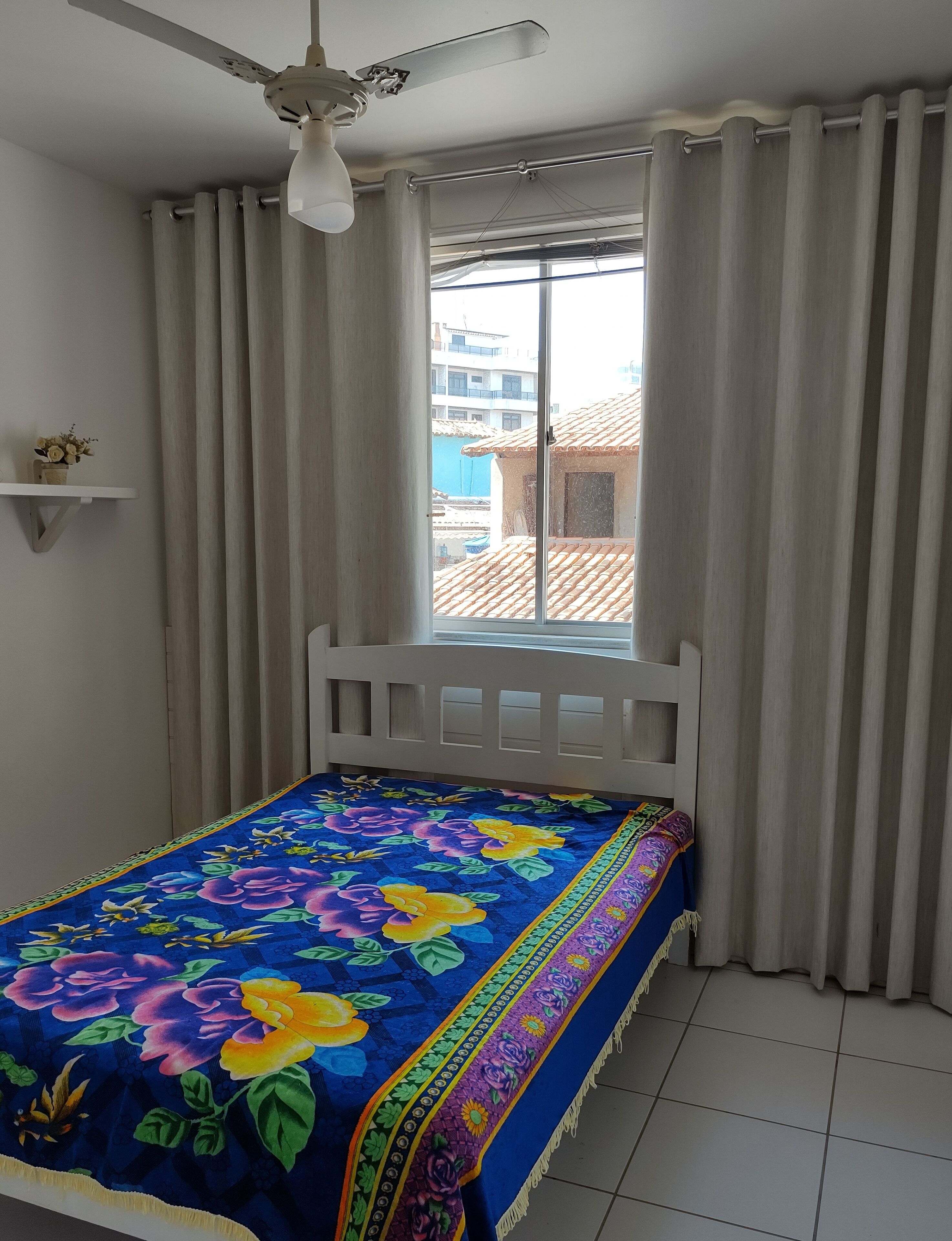 A bedroom with a bed, a ceiling fan, a window with curtains, and a view of buildings outside.