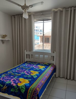 Quarto com cama, ventilador de teto, janela com cortinas e vista para prédios.