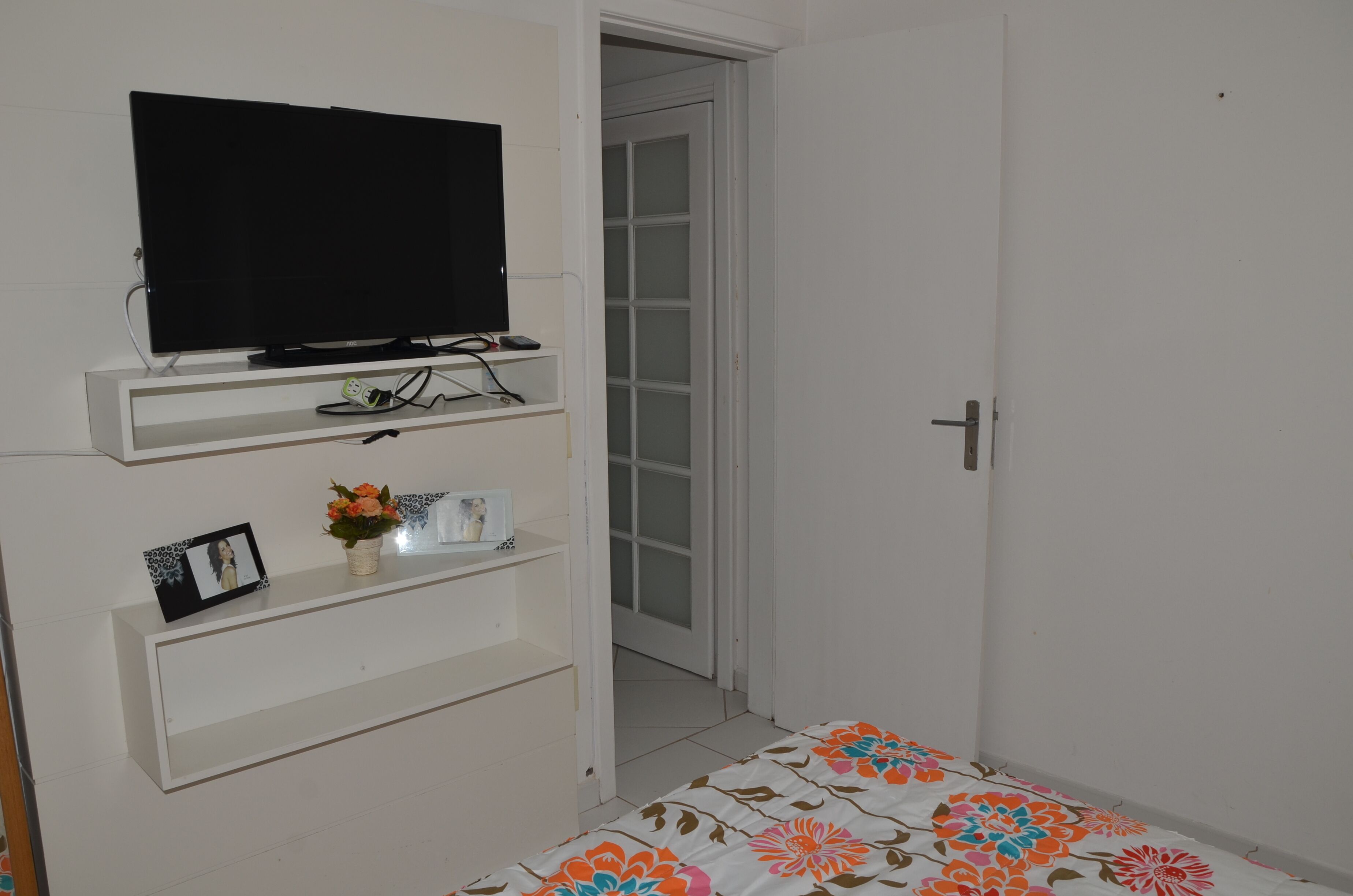 Ein Zimmer mit einem Fernseher auf einem Schrank, einer kleinen Ablage mit Bilderrahmen und einer Vase, und einer Tür zu einem anderen Raum.