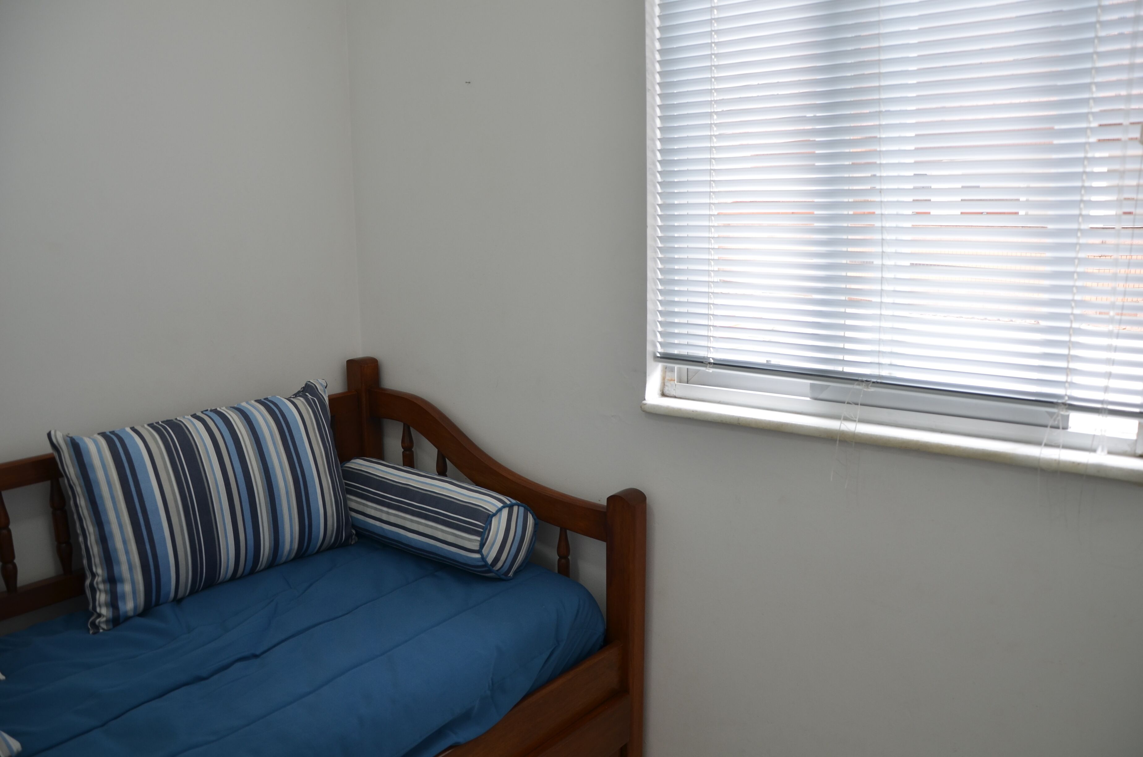 Ein Einzelbett mit Holz Kopfteil, zwei gestreifte Kissen und einer blauen Bettdecke. Rechts ist ein Fenster mit Jalousien zu sehen.