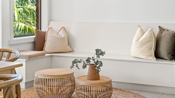 A cozy seating area with a cushioned bench, two round woven side tables, and a vase with greenery.