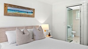 A hotel room with a bed, pillows, a nightstand with a lamp, a painting on the wall, and a bathroom visible through an open door.