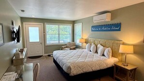 Ein Schlafzimmer mit Bett, Nachttischen, Schreibtisch und Stuhl. Es gibt ein Fenster mit Blick, eine Tür und ein Schild an der Wand.