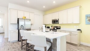 A modern kitchen with white cabinetry, a central island with bar stools, a stainless steel refrigerator, and a microwave.