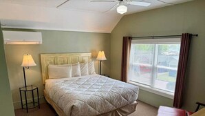 Un dormitorio con cama, dos lámparas de noche, ventilador de techo, aire acondicionado, ventana con cortinas y una pequeña mesa de madera.