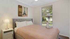 Ein Schlafzimmer mit einem Bett, einem Nachttisch, einer Lampe, einem Fenster mit Jalousien und einem Gemälde vom Eiffelturm.
