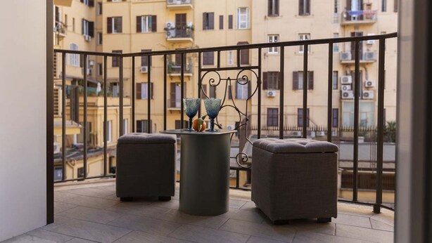 A balcony with two grey ottomans and a round table with decorative items, overlooking a residential area with yellow buildings. - The bright, spacious apartment is located in Via Luigi Settembrini, on the 1st floor of an elegant Art Nouveau residential building with porter's lodge and elevator, from the early 20th century. During the most recent renovation work, some antique, elegan (Roma)