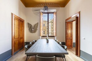 Un espace repas comprenant une longue table, des chaises, un luminaire suspendu et une vue sur les bâtiments à travers une fenêtre.
