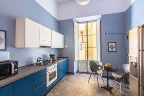 Een kleine keuken met blauwe kastjes, een magnetron en een eethoek met een tafel en stoelen.
