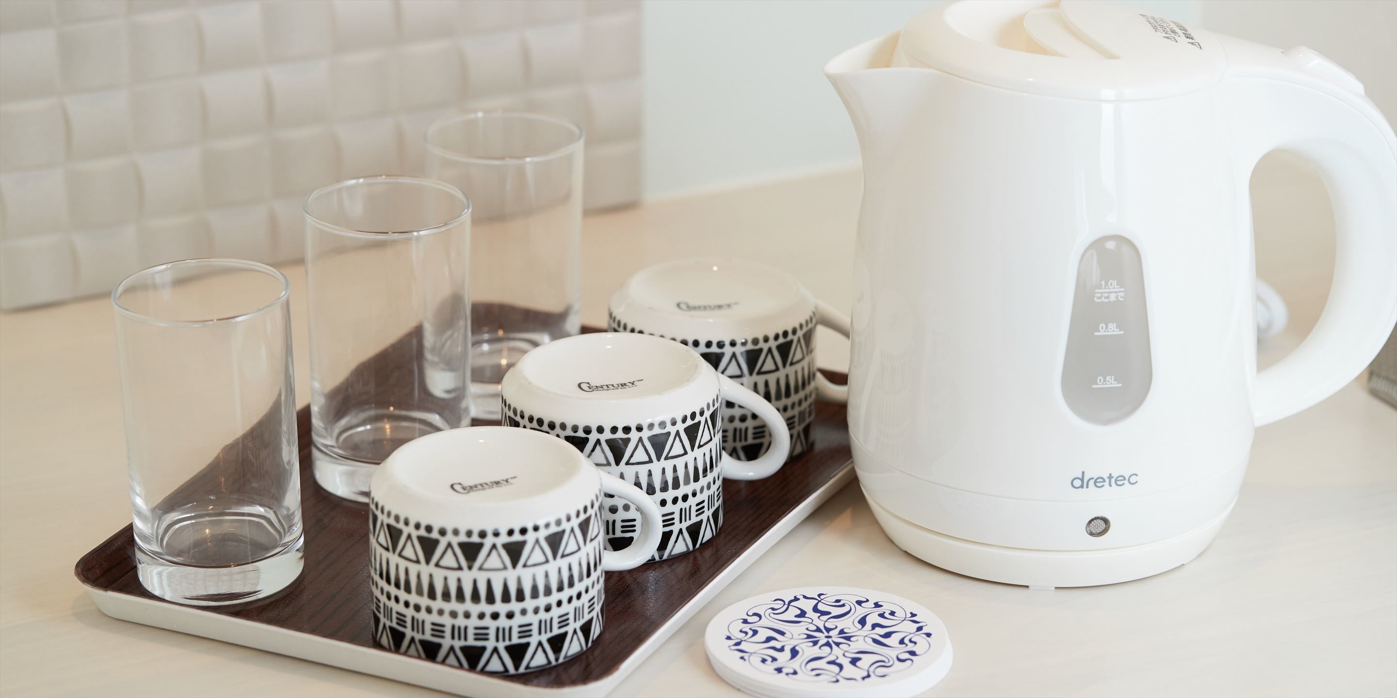 Una tetera eléctrica blanca, cuatro tazas con diseños y dos vasos transparentes sobre una bandeja.