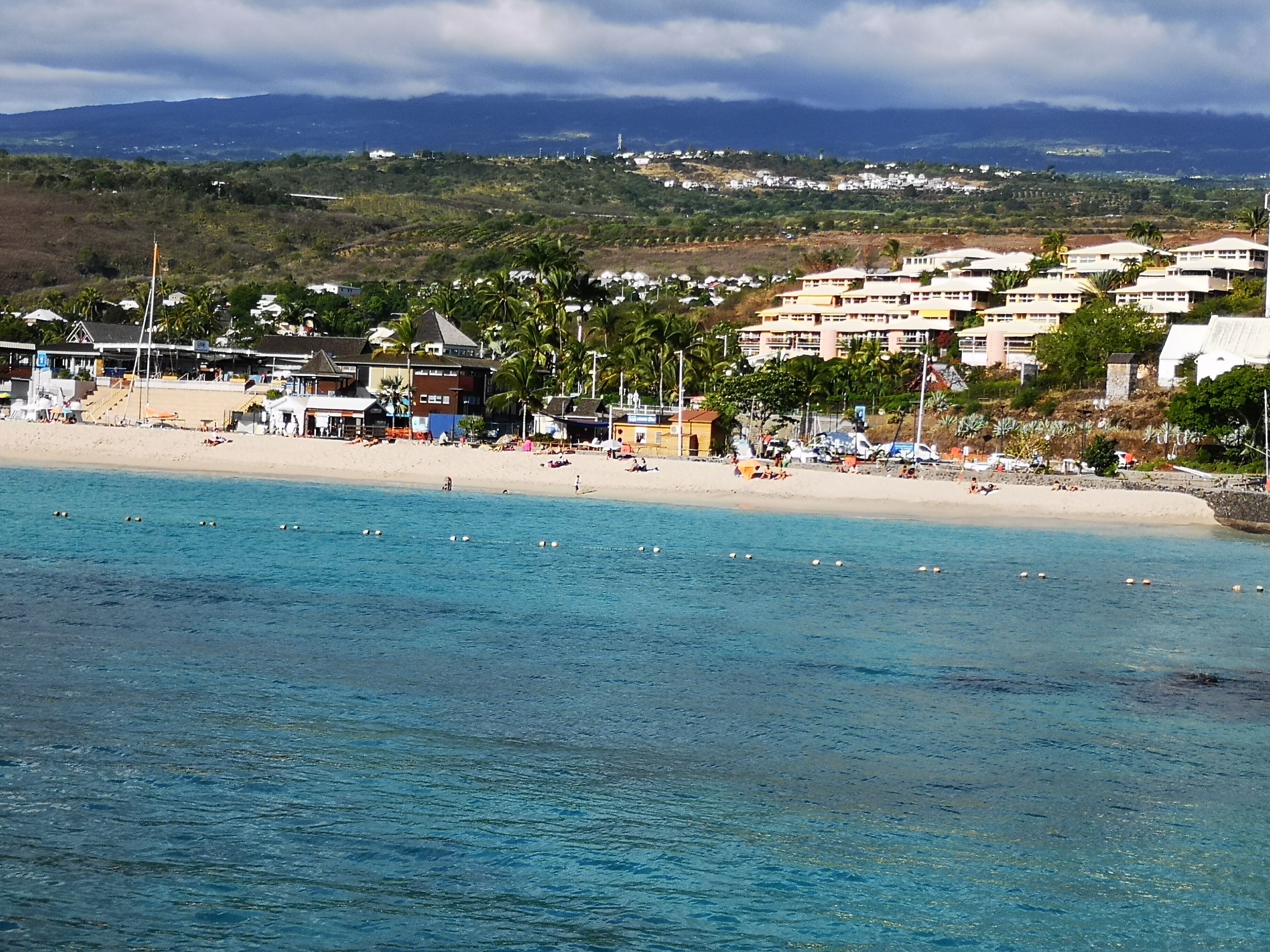 Escena costera de playa con aguas azul turquesa, una orilla arenosa y edificios al fondo.