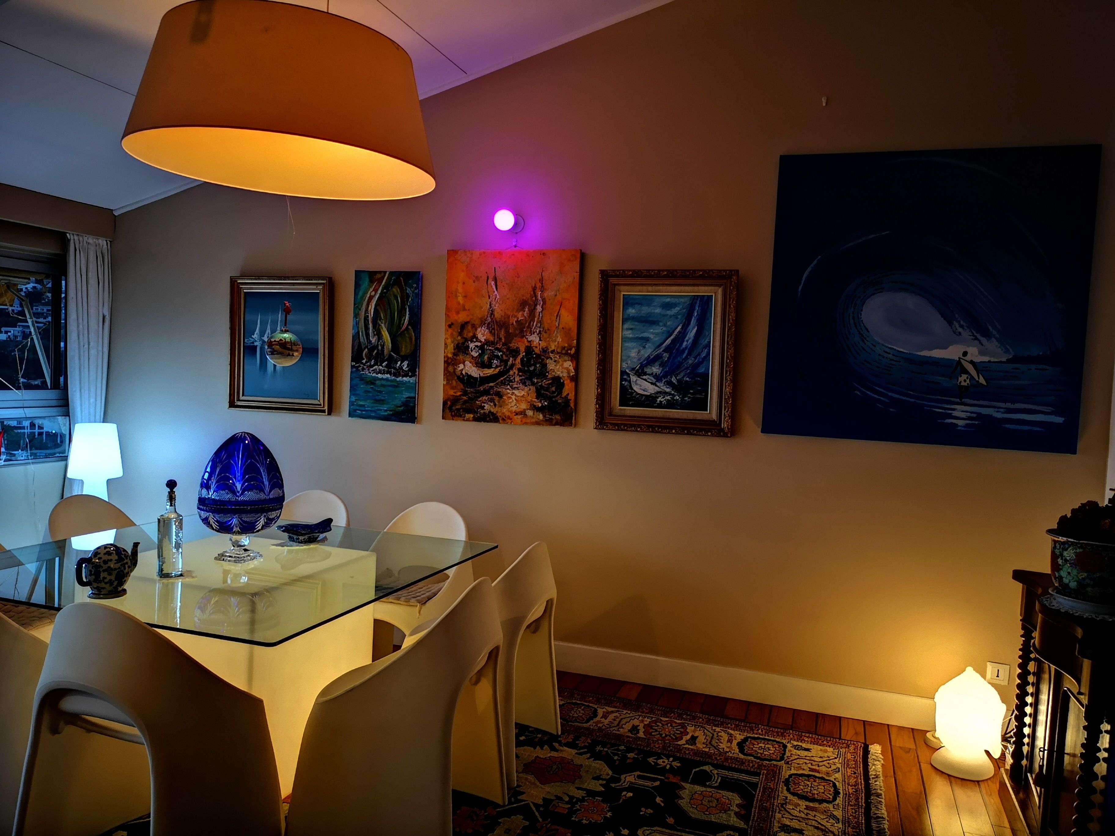 A dining area with a glass table, chairs, and a hanging lamp. There are framed artworks on the wall and a rug on the floor.