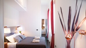 Camera d'albergo con un letto, lampade da comodino, un tavolino, un vaso con decorazioni e vista sul corridoio.