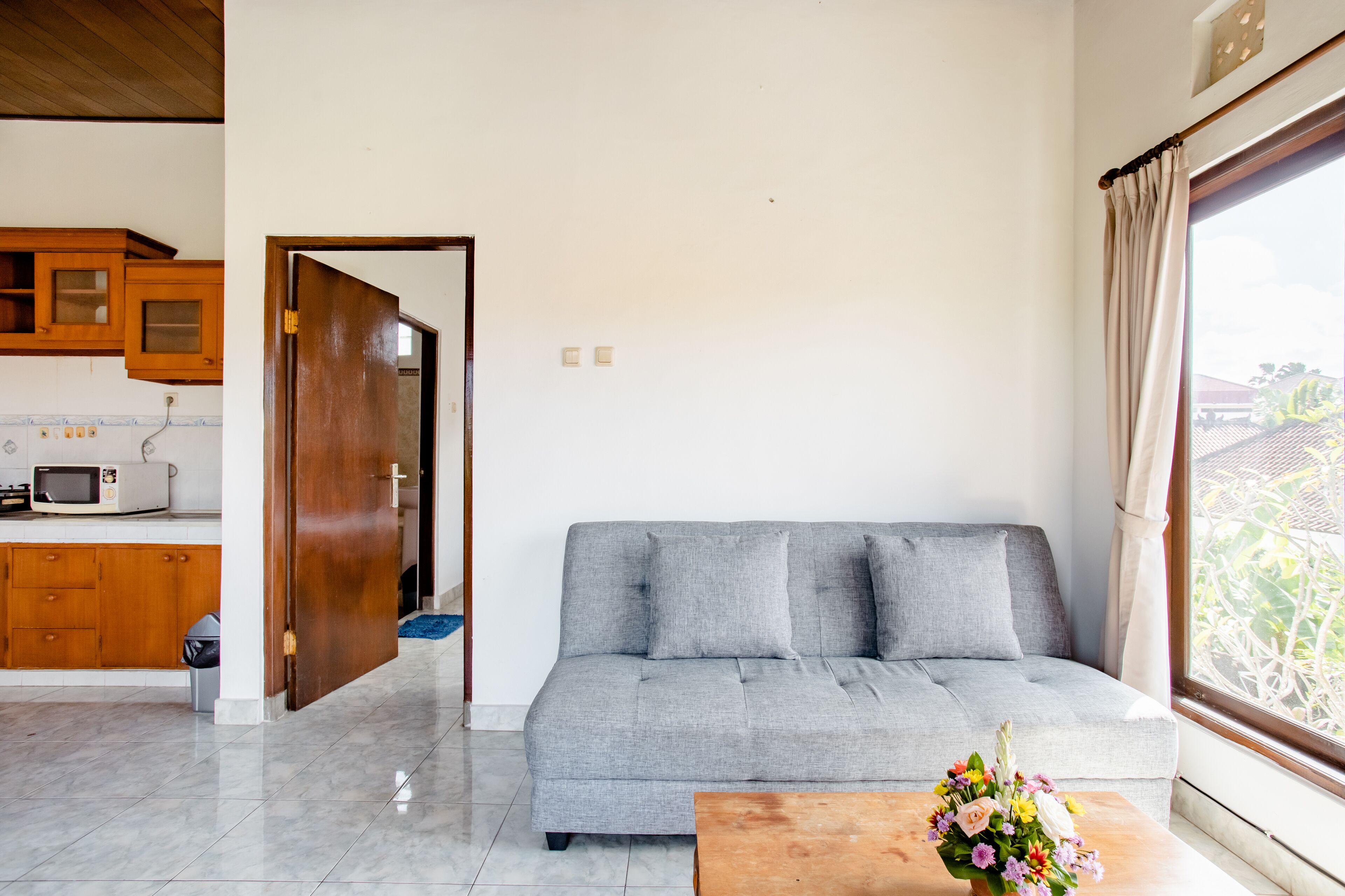 Una sala de estar con un sofá gris, armarios de madera, un microondas y una ventana con cortinas.