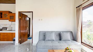 Una sala de estar con un sofá gris, armarios de madera, un microondas y una ventana con cortinas.