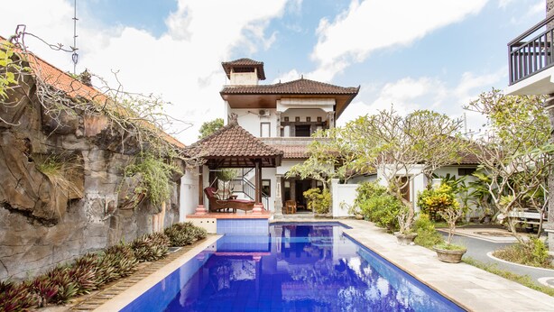 A swimming pool with a tiled deck, a covered seating area, and a building with a tiled roof in the background. - Rumah Akira Seminyak (Seminyak)