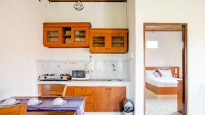 A compact kitchen with wooden cabinets, a microwave, and a gas stove. There is a dining table with chairs and a small trash bin. A bedroom with a bed and a nightstand is visible through an open doorway. - Rumah Akira Seminyak (Seminyak)