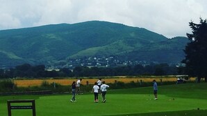 A golf course with a wooden bench and a sign, surrounded by green hills and a village in the background.