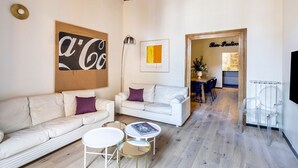 Un soggiorno moderno con un divano, due tavolini e un tavolino da caffè. Appeso al muro, si nota un'opera d'arte incorniciata e una lampada. Un'apertura conduce a una zona cucina.