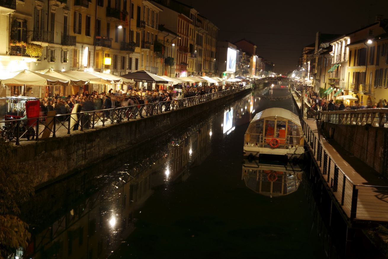 A canal-side walkway with buildings on either side, a boat docked on the right, and people walking along the path.