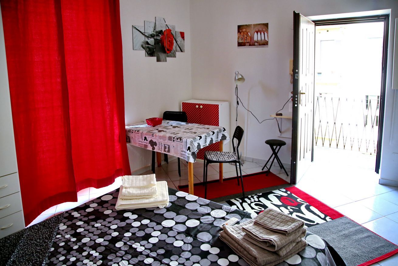 A room with a bed, a table with a polka dot tablecloth, a chair, a red curtain, and a door leading outside.
