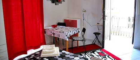 Una habitación con una cama, una mesa con mantel de lunares, una silla, una cortina roja y una puerta que da al exterior.
