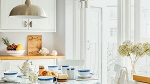 A dining area with a table set for two, featuring blue and white dinnerware, a white teapot, and a wooden board with fruits and a menu.