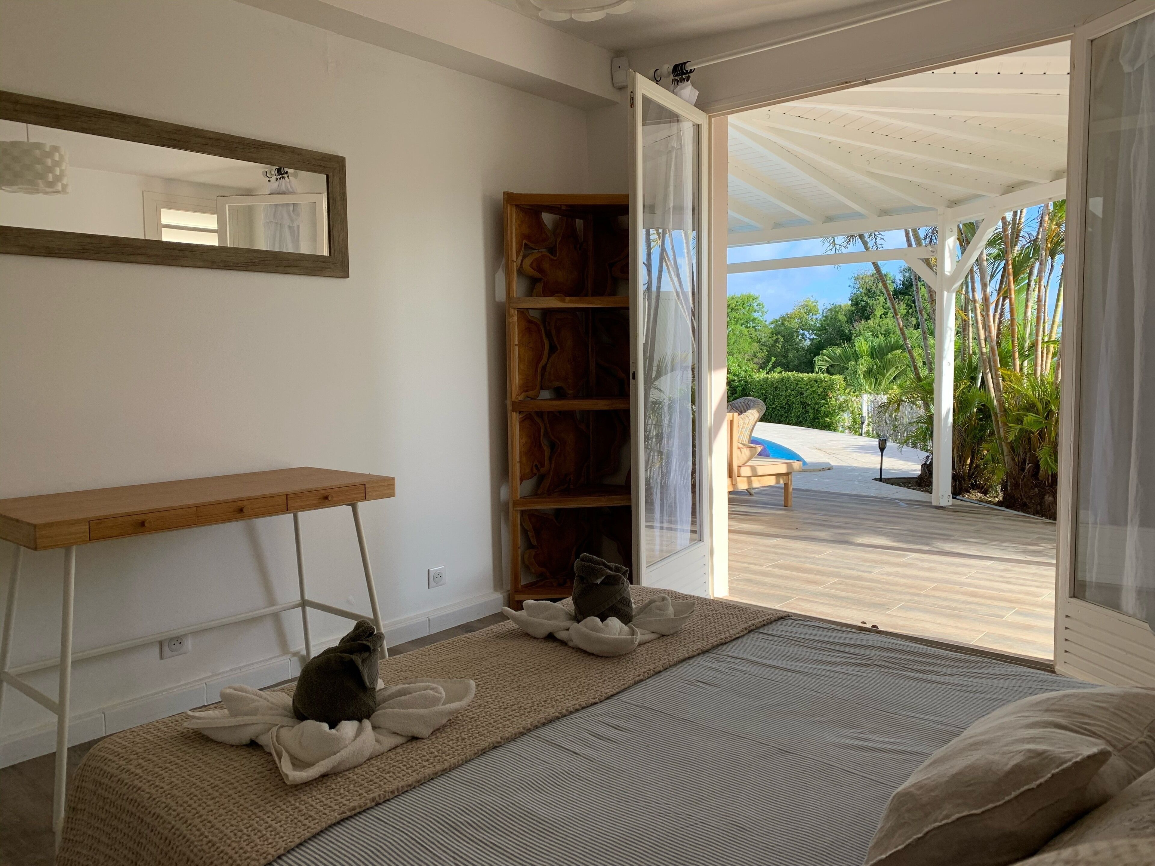 Une pièce dotée d’un bureau en bois, d’un grand miroir et d’une porte-fenêtre coulissante donnant sur une terrasse extérieure avec une piscine et un fauteuil de relaxation.