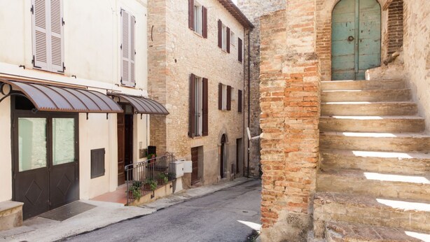 A narrow street with buildings on either side, featuring a mix of brick and plaster facades, a metal awning, and a green door. - Medieval village flat, second floor with Wi-Fi & aircon (Compignano)