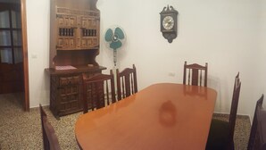 Uma área de refeições com uma mesa e cadeiras de madeira, um relógio na parede e um ventilador.