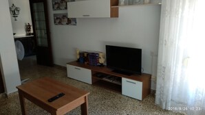 Uma sala de estar com uma mesa de centro de madeira, uma TV de tela plana em um suporte de madeira e uma pequena estante com objetos decorativos.