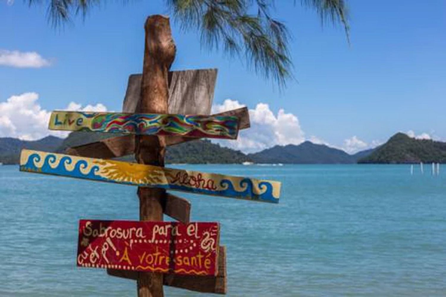 A wooden signpost with multiple directional signs against a backdrop of a clear blue sea and distant mountains.