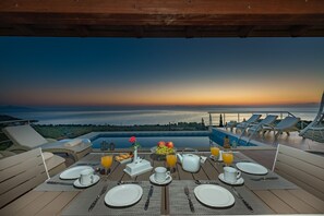 Ein Esstisch ist für das Frühstück gedeckt, mit Blick auf den Sonnenuntergang über dem Wasser, Liegestühle und ein Poolbereich.